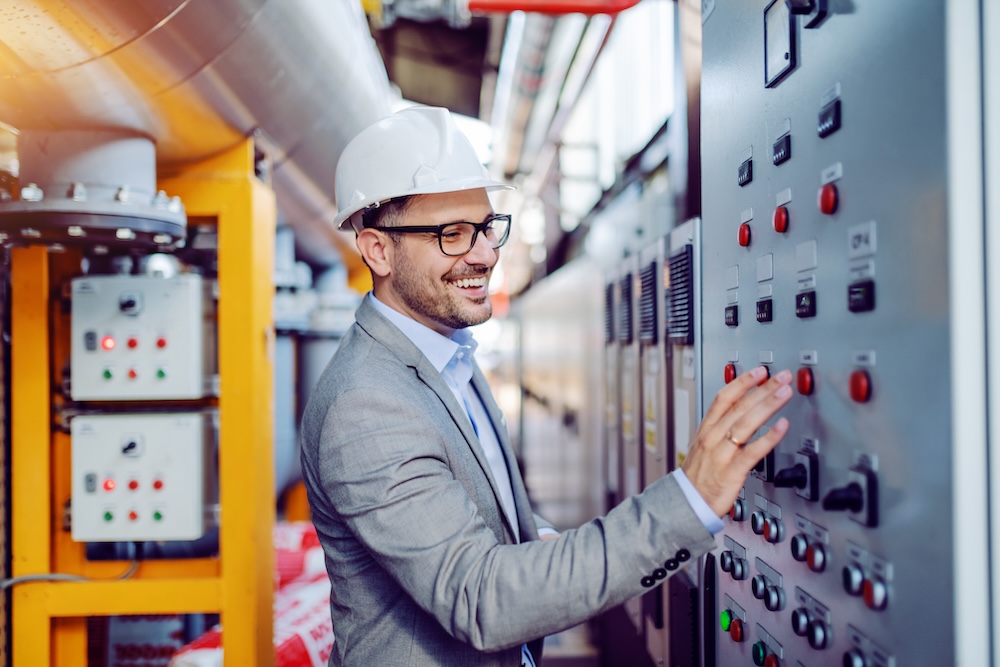 Smiling engineer in gray suit and with helmet on head turning switch on inside a power plant.